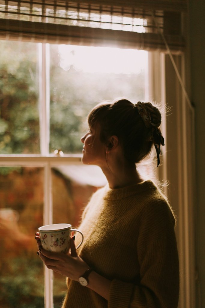 Woman holding a cup of coffee, looking out of a window in morning light